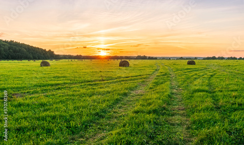 Scenic view at beautiful sunset in green shiny field with hay stacks, bright cloudy sky, country road and golden sun rays with glow, summer valley landscape