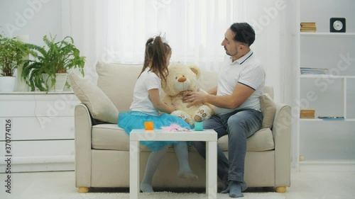 Cute girl and her father sitting on the sofa, hugging teddy bear. Father is touching daughter's nose, then teddy's. photo