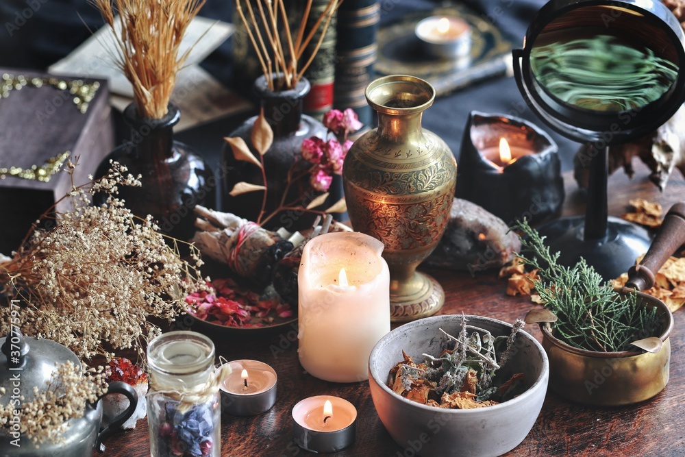 Dried plants, herbs and yellow rose flower petals in a grey, dirty clay pot. Wiccan witch altar with ingredients on it ready to make cast a spell. Evergreens, books, burning candles in dark background