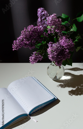 Still life of bright branch of lilac in the glass vase with open diary on white table on grey curtains background