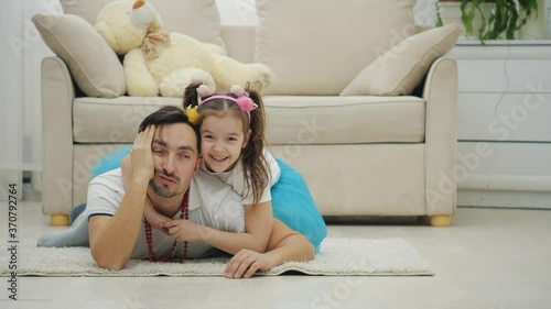 Pretty little girl with pigtails is smiling, hugging her father who is lying on the floor, tired and sleepy. photo