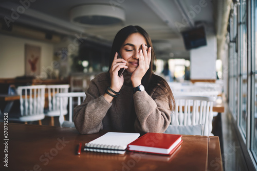 Smiling brunette female student feeling happy talking on phone with friend sitting in cafe interior, young journalist satisfied with successful project getting offer having conversation via smartphone