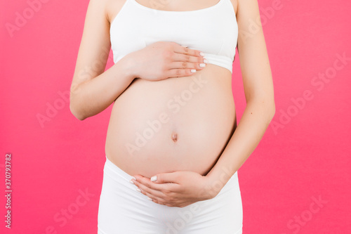 Cropped image of pregnant woman wearing white underwear. Young mother is hugging her belly expecting a baby at pink background. Copy space