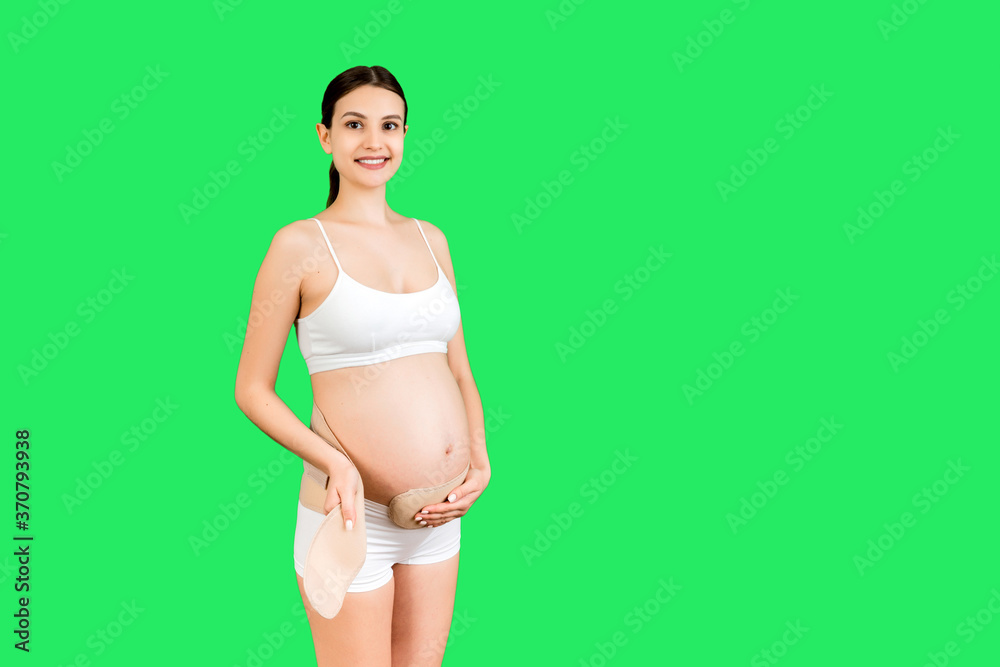 Portrait of pregnant woman in underwear putting on pregnancy bandage at  green background with copy space. Orthopedic abdominal support belt concept  Stock Photo