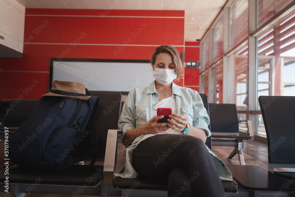 Young woman traveler in a protective medical mask with a smartphone in her hands is waiting for her flight at the airport, a new normal lifestyle Young woman traveler in a protective medical mask with a smartphone in her hands is waiting for her flight at the airport, a new normal lifestyle