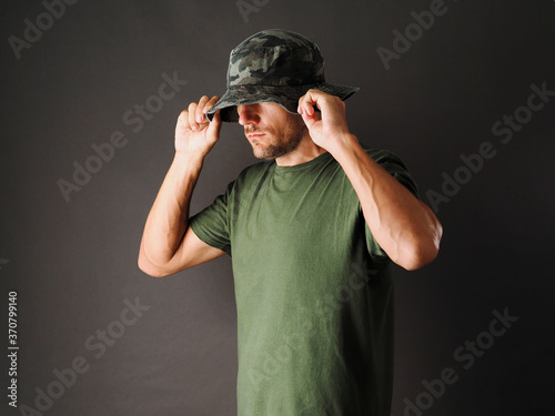 The man is wearing a green camouflage Panama hat and a green t-shirt on a grey background.