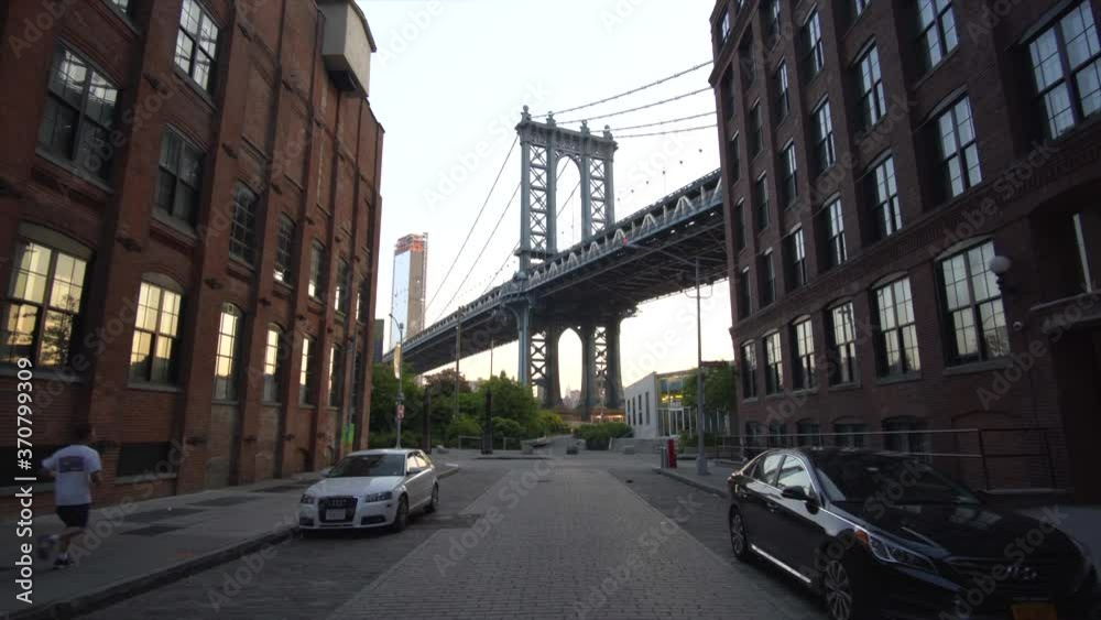 Brooklyn Bridge View from Dumbo with dumbo apartment houses and parked cars on the side in Summer in early morning light with Runner passing by Brooklyn Bridge View from Dumbo with dumbo apartment houses and parked cars on the side in Summer in early morning light with Runner passing by