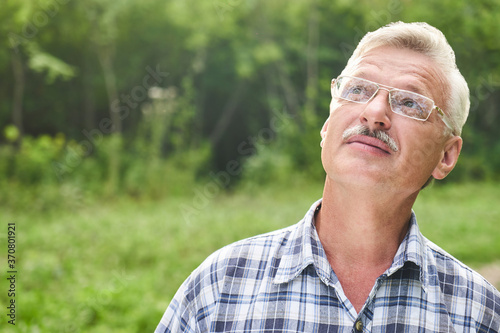 Close-up portrait of handsome gray-haired adult man with mustache and glasses smiling on the background of a summer forest park