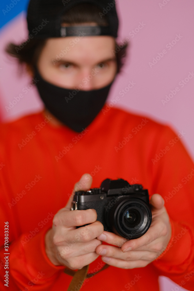 Male photographer. A young man of 25-30 years old in a black protective mask and in a red sweatshirt holds a mirrorless camera in his hands. 