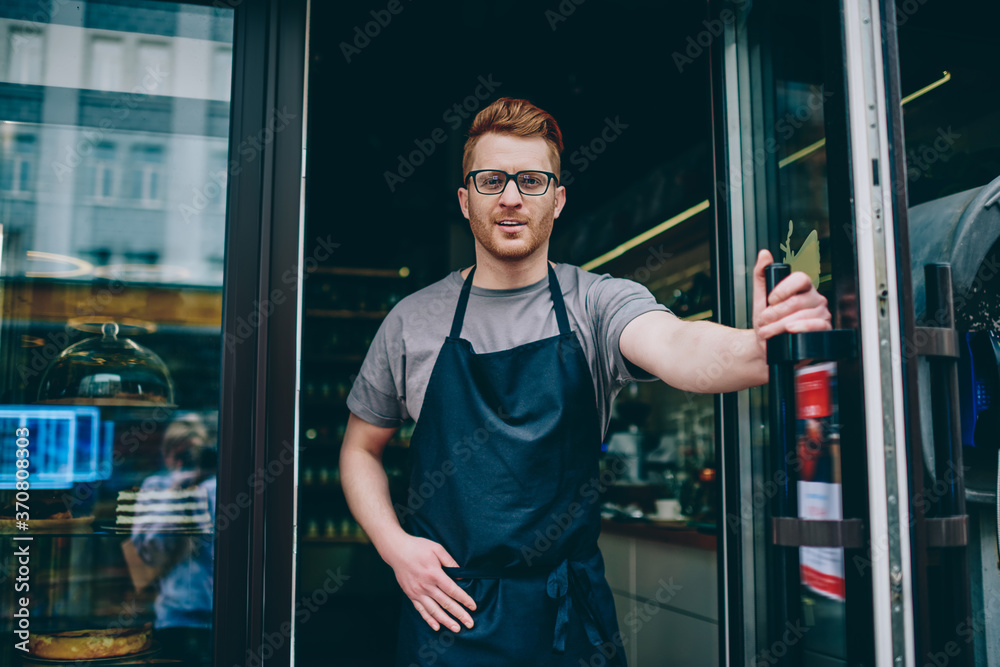 Portrait of successful caucasian barista in apron smiling at camera and inviting guests in coffee shop.Cheerful male waiter smiling at camera while standing at entrance to own cafeteria Portrait of successful caucasian barista in apron smiling at camera and inviting guests in coffee shop.Cheerful male waiter smiling at camera while standing at entrance to own cafeteria