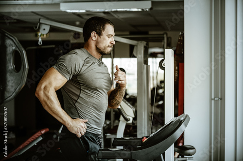 Fitness in gym, sport and healthy lifestyle concept. Handsome athletic man in grey shirt runs on treadmill. Bodybuilder male model with trained body does cardio workout