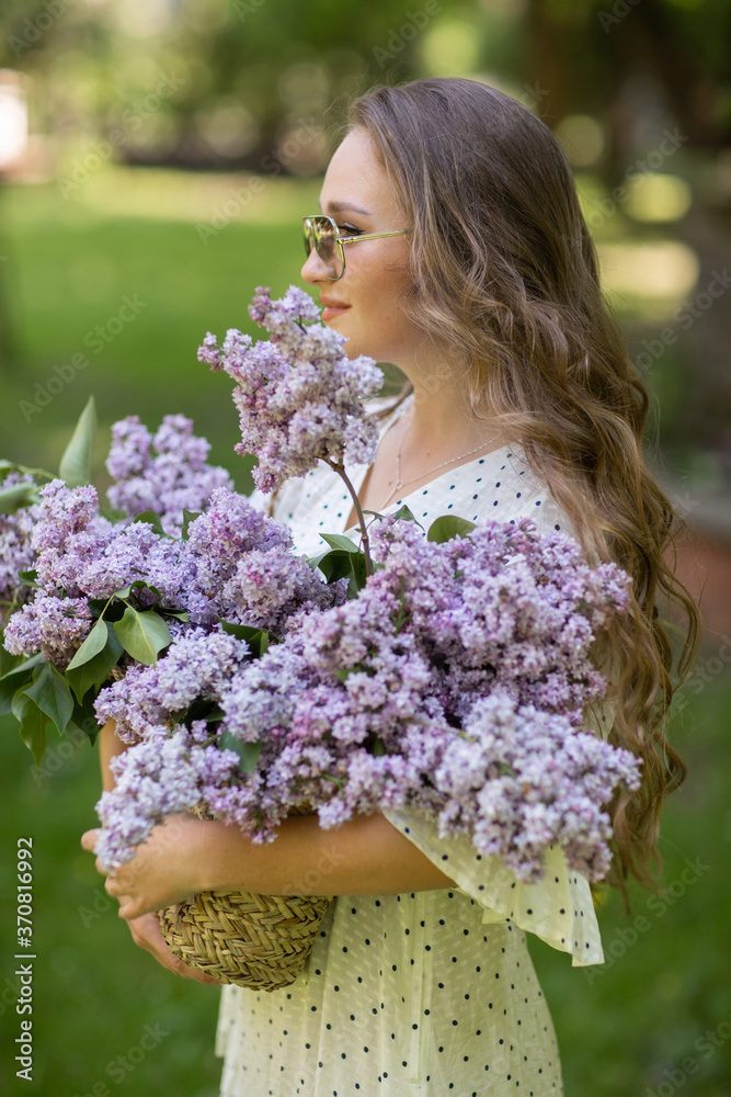 The girl in white dress and sunglasses holds in her hands a wicker basket with flowers. Basket with lilacs. Girl and flowers. Walk with a basket of lilacs in the hands. Floristics The girl in white dress and sunglasses holds in her hands a wicker basket with flowers. Basket with lilacs. Girl and flowers. Walk with a basket of lilacs in the hands. Floristics