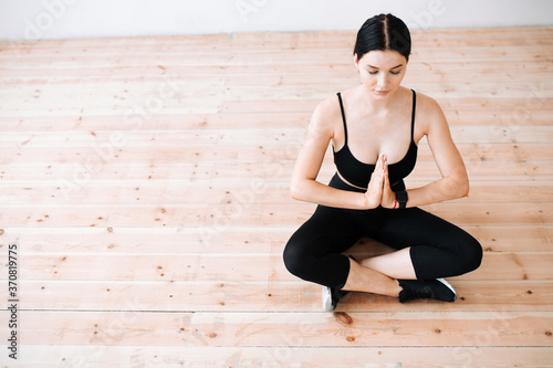 Asian woman sits in lotus position on the wooden floor, joins her hands in front of her and meditates