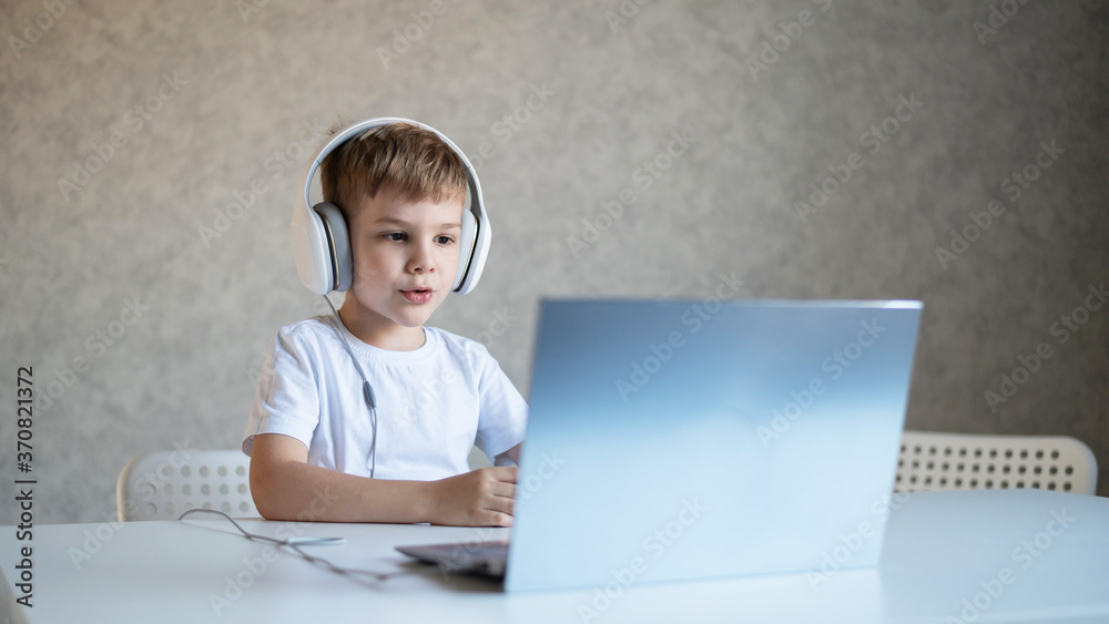 Cute little boy is watching cartoons or a movie on a laptop. Caucasian child sits at a table at home and listens to music on headphones. Cute little boy is watching cartoons or a movie on a laptop. Caucasian child sits at a table at home and listens to music on headphones.