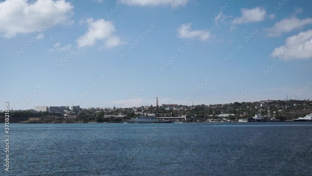 A gray warship sails in a Mediterranean bay against the backdrop of a small coastal town. Footage. Beautiful seascape with a large patrol ship on a sunny day and blue water in calm A gray warship sails in a Mediterranean bay against the backdrop of a small coastal town. Footage. Beautiful seascape with a large patrol ship on a sunny day and blue water in calm