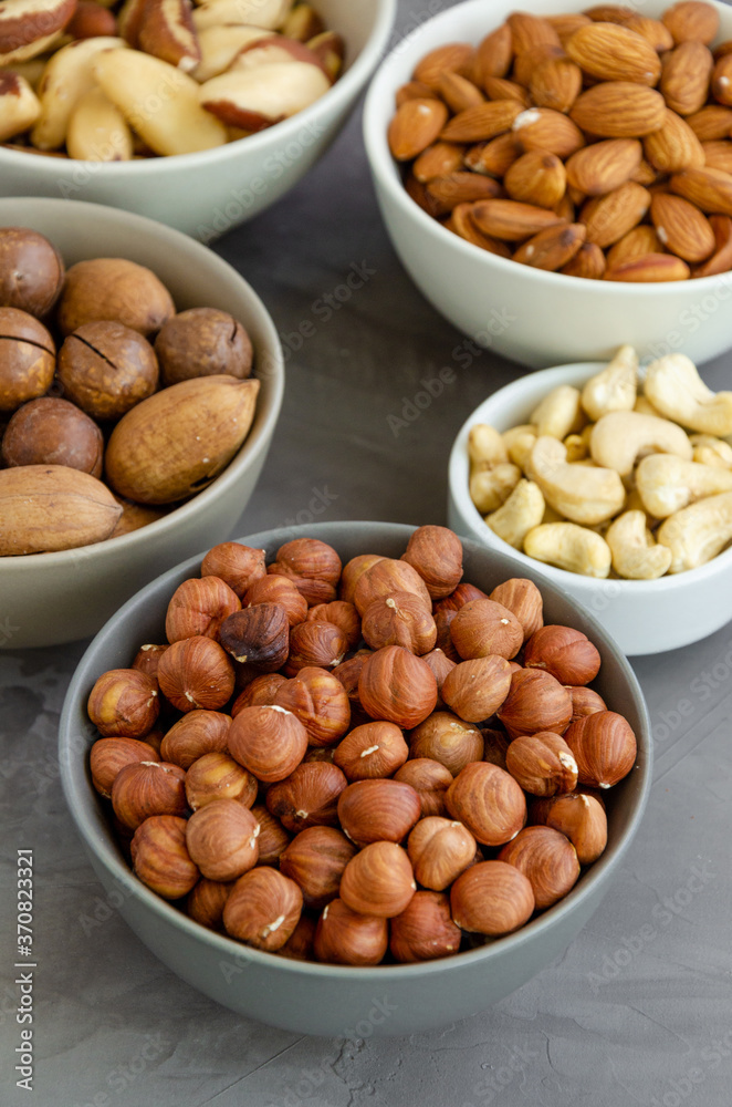 Hazelnuts, almonds, Brazilian nuts, cashews, macadamia, pecans and pistachios in bowls on a dark concrete background. Healthy food. Vertical, copy space. Hazelnuts, almonds, Brazilian nuts, cashews, macadamia, pecans and pistachios in bowls on a dark concrete background. Healthy food. Vertical, copy space.