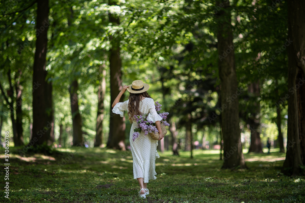 Woman with long hair and straw hat. The girl holds in her hands a wicker basket with flowers. Basket with lilacs. Woman and flowers. Walk with a basket of lilacs in the hands. Floristics.