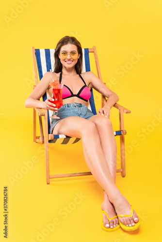  woman in sunglasses and flip flops sitting on deck chair and holding cocktail on yellow