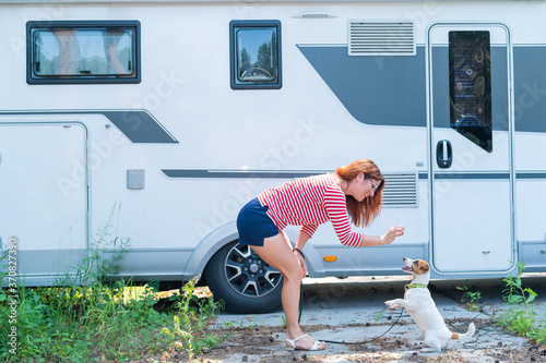Beautiful caucasian woman is training a dog next to the motorhome. Travel with your pet in a mobile RV. Red-haired girl playing with Jack Russell Terrier. Female trailer driver.