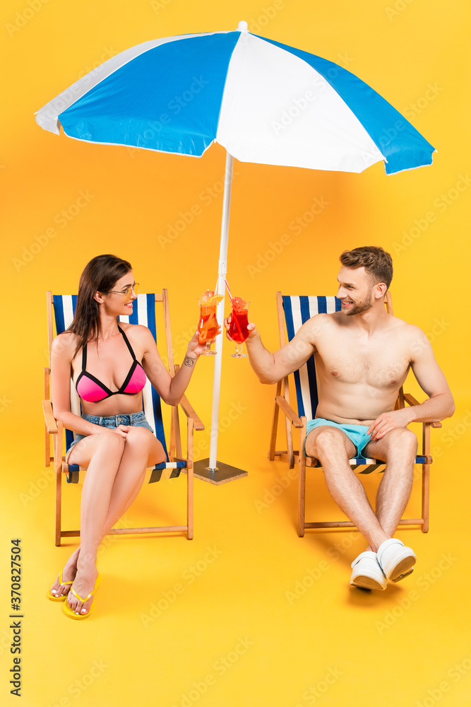  couple clinking cocktails and sitting on deck chairs near beach umbrella on yellow