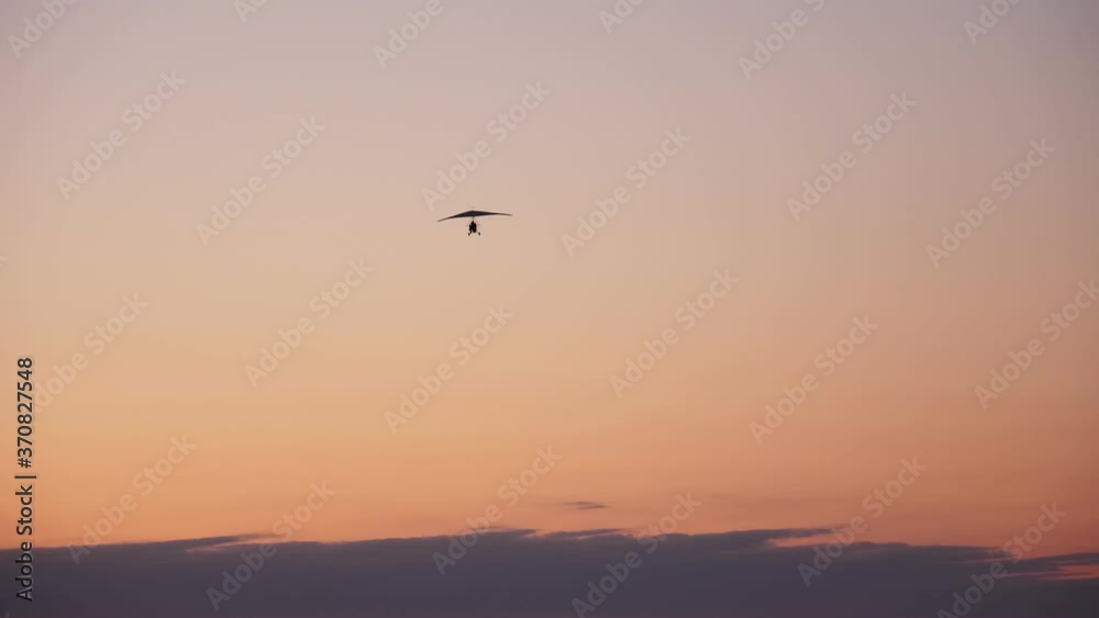 dark silhouette of motorized hang glider with passenger and pilot, flies, soars in the air, against the background of a bright orange sunset dark silhouette of motorized hang glider with passenger and pilot, flies, soars in the air, against the background of a bright orange sunset