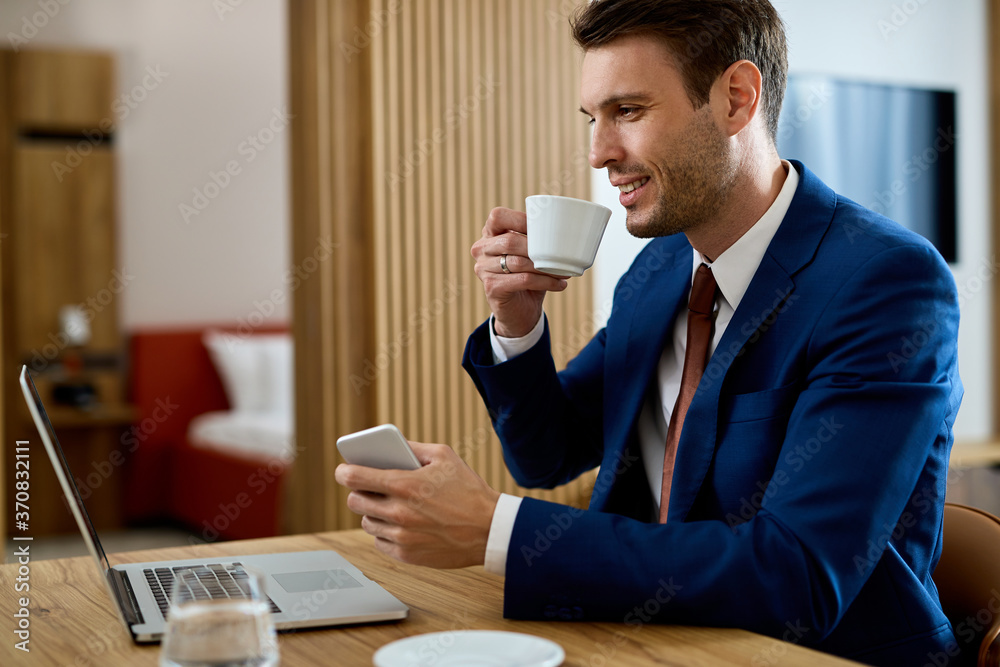 Happy businessman having a cup of coffee while reading e-mail on laptop in hotel room. Happy businessman having a cup of coffee while reading e-mail on laptop in hotel room.
