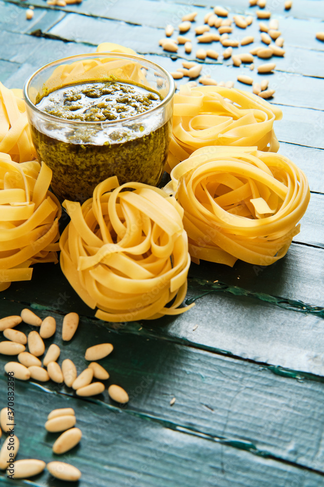 From above of tasty pesto salsa with olive oil surrounded by uncooked pasta rolls and spilled crunchy pine nuts on wooden table From above of tasty pesto salsa with olive oil surrounded by uncooked pasta rolls and spilled crunchy pine nuts on wooden table