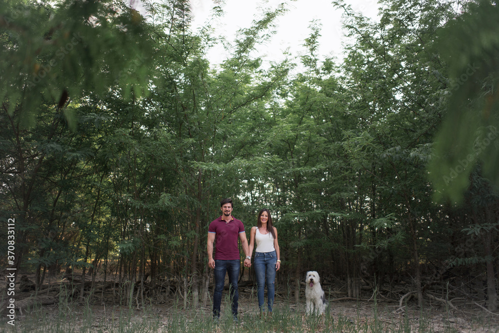 Side view of tender couple holding hands while standing with Old English Sheepdog in garden on sunny day looking at camera Side view of tender couple holding hands while standing with Old English Sheepdog in garden on sunny day looking at camera