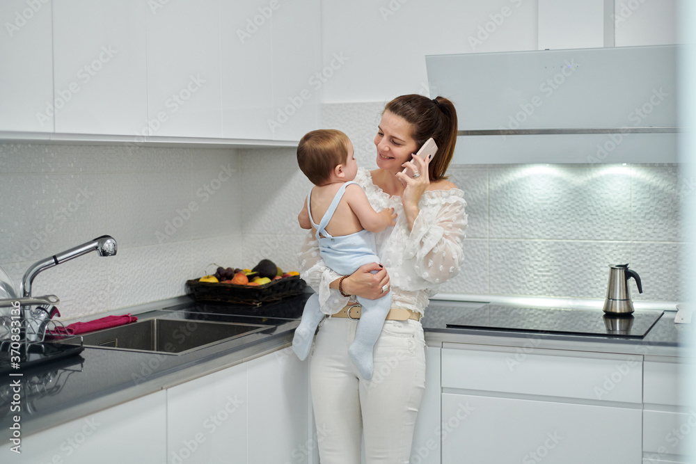 Young woman smiling at cute little baby while standing in kitchen and talking on mobile phone at home Young woman smiling at cute little baby while standing in kitchen and talking on mobile phone at home
