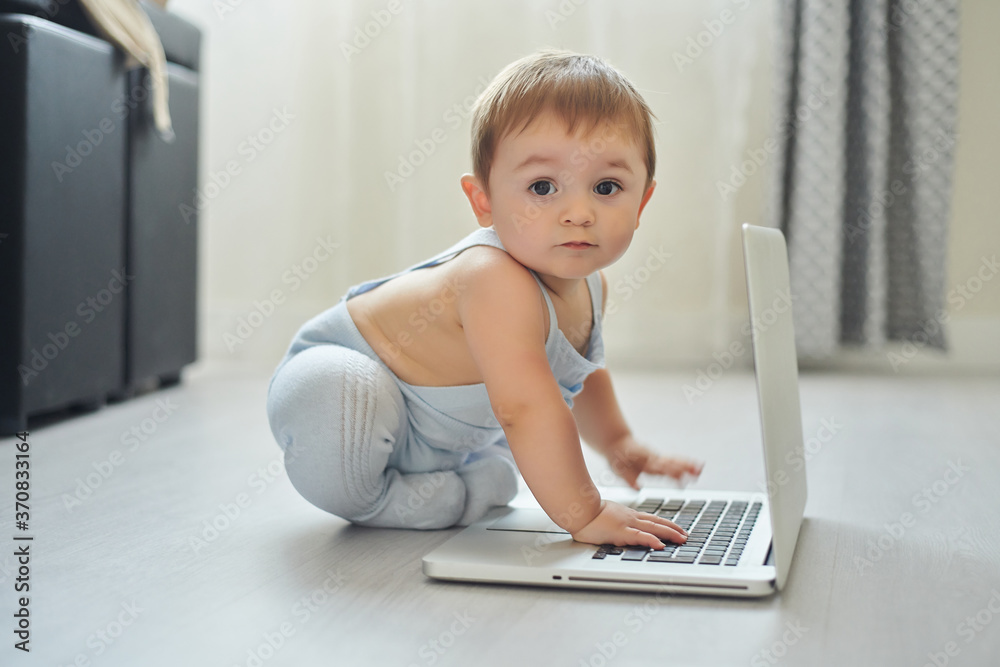 Side view of happy little toddler sitting on floor and pressing keys on laptop while playing alone in light room at home Side view of happy little toddler sitting on floor and pressing keys on laptop while playing alone in light room at home
