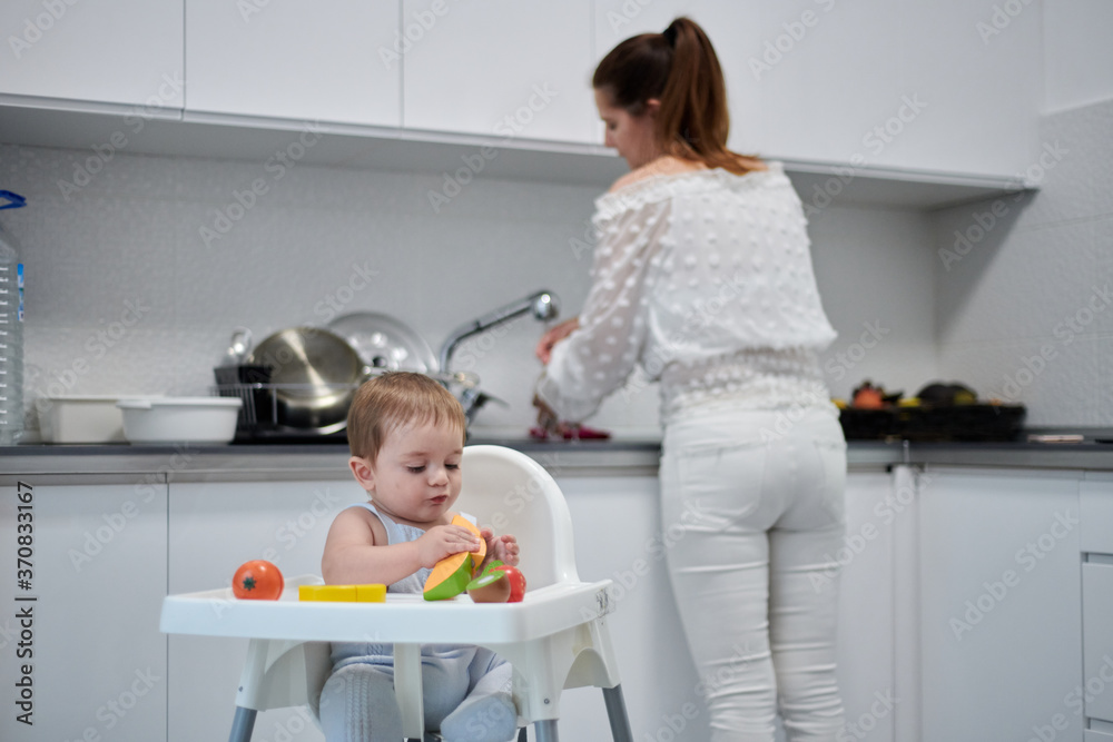 Focused little baby sitting in high chair with various toy fruits on table while mother standing near sink and washing grapes in cozy light kitchen Focused little baby sitting in high chair with various toy fruits on table while mother standing near sink and washing grapes in cozy light kitchen
