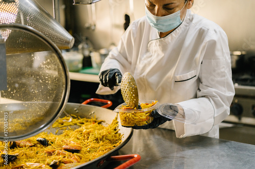 Focused female chef placing delicious noodle with seafood in plastic container for delivery while working in restaurant kitchen during coronavirus pandemic