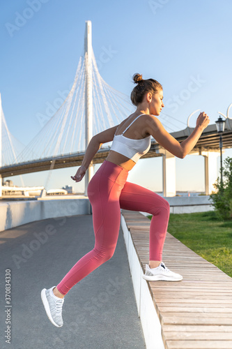 Fitness woman practicing jumping exercises on wooden bench outdoor. Side view of sportswoman wearing pink leggings, making cardio workout, bridge on background.  © DimaBerlin