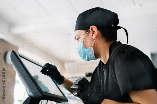 Side view serious female cashier wearing gloves and mask tapping on cash register touchscreen during duty in contemporary cafe