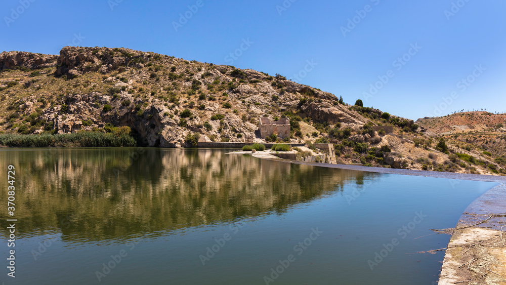 The dam of the Elche reservoir in eastern Spain in sunshine and blue skies. The dam has an overflow. In the background are mountains.