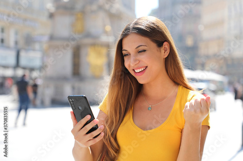 Happy excited satisfied woman holding smart phone reading good news outdoor and celebrating in city street