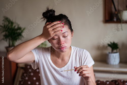 Exhausted ethnic female sitting on bed at home and measuring temperature with thermometer while having cold and looking away photo