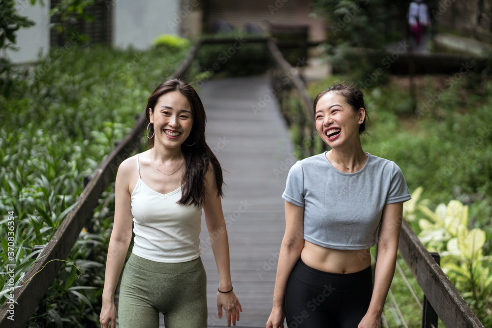 Smiling fit Asian girlfriends wearing tight casual clothes strolling along narrow wooden bridge in countryside and chatting Smiling fit Asian girlfriends wearing tight casual clothes strolling along narrow wooden bridge in countryside and chatting