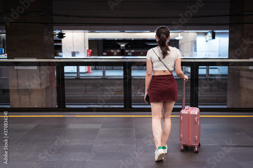 Back view of unrecognizable young female in mini skirt walking with suitcase in railway station while waiting for train