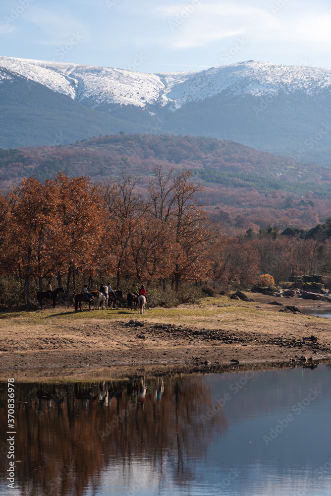Company of horsemen riding horses along shore of calm river in autumn in mountainous area on sunny day Company of horsemen riding horses along shore of calm river in autumn in mountainous area on sunny day
