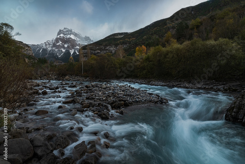 Amazing natural landscape of rapid shallow river streaming among boulders in mountainous terrain with forest and rocky peak in background in cloudy autumn day