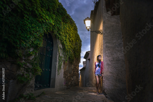 Calm young ethnic female in casual colorful clothes leaning on stone wall of aged building while exploring ancient street of Tossa de Mar municipality in Spain in summer evening photo