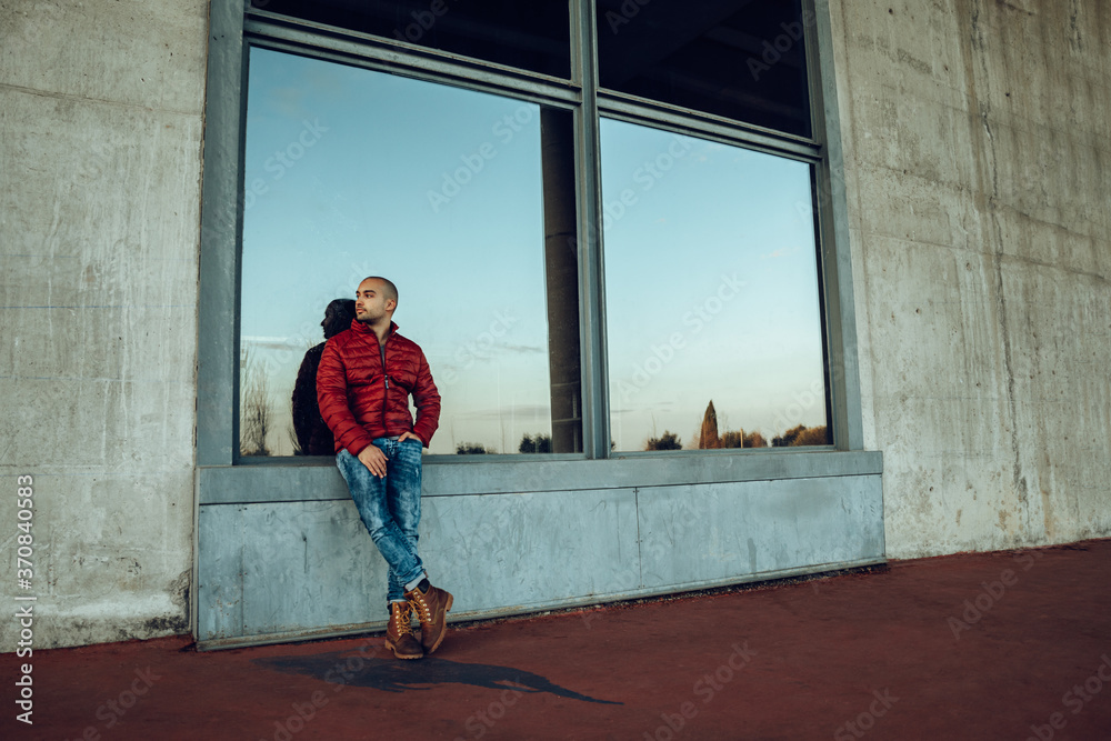 Confident young unshaven male in red warm jacket and jeans looking away while standing and leaning against a big window Confident young unshaven male in red warm jacket and jeans looking away while standing and leaning against a big window