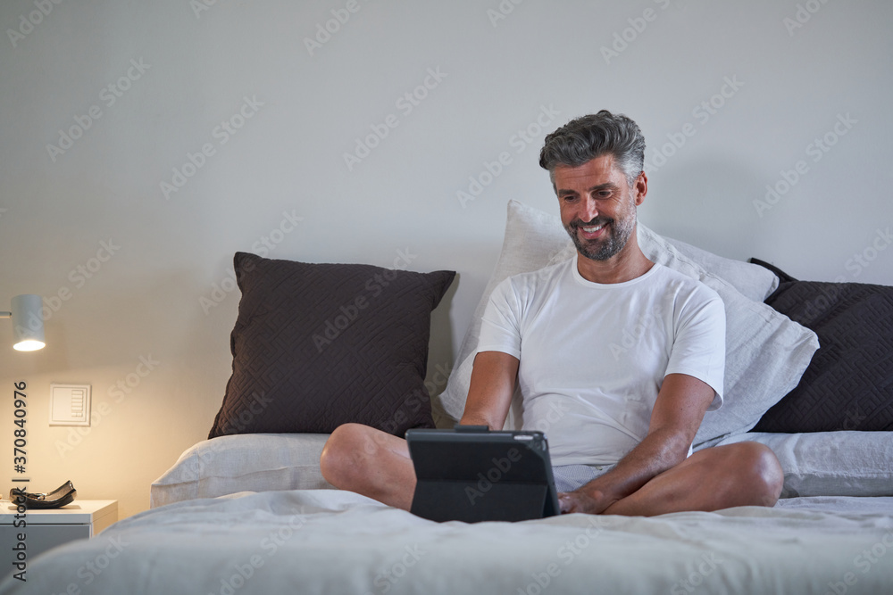 Smiling middle aged bearded male in casual clothes sitting on bed and browsing laptop while resting in cozy bedroom in evening Smiling middle aged bearded male in casual clothes sitting on bed and browsing laptop while resting in cozy bedroom in evening