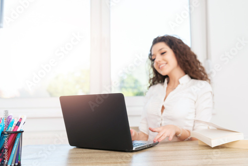Confident woman with curly brown hair sitting in office by the window  using laptop and smiling.