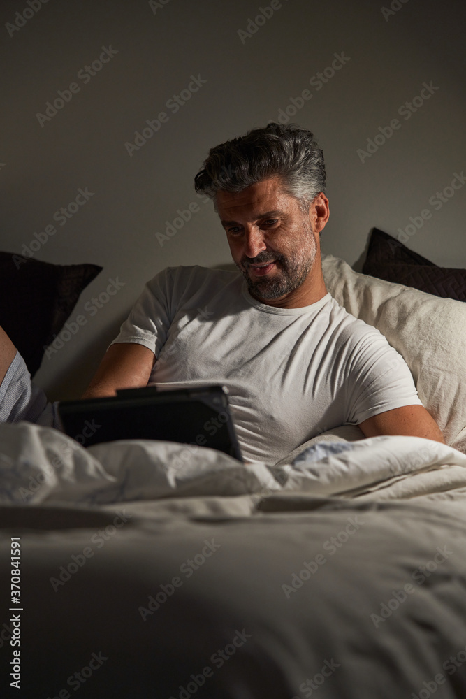 Smiling middle aged bearded male in casual clothes lying on bed and browsing laptop while resting in cozy bedroom in evening Smiling middle aged bearded male in casual clothes lying on bed and browsing laptop while resting in cozy bedroom in evening