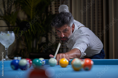 Confident focused gray haired male in white shirt preparing for shot with cue while playing billiards game