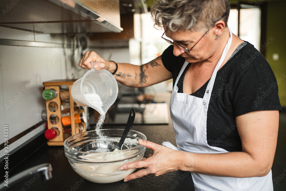 Crop housewife wearing apron standing at counter in kitchen and pouring sugar in glass bowl while preparing dough for pastry Crop housewife wearing apron standing at counter in kitchen and pouring sugar in glass bowl while preparing dough for pastry