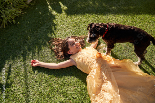 High angle of happy little girl in beautiful dress lying on green grass and laughing while playing with fluffy dog during summer day in garden