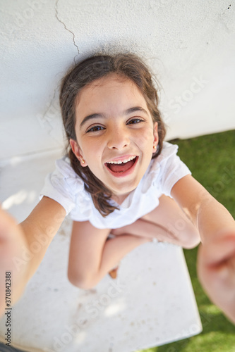 Adorable school aged girl in casual clothes smiling and looking at camera while sitting near wall of house in sunny summer day